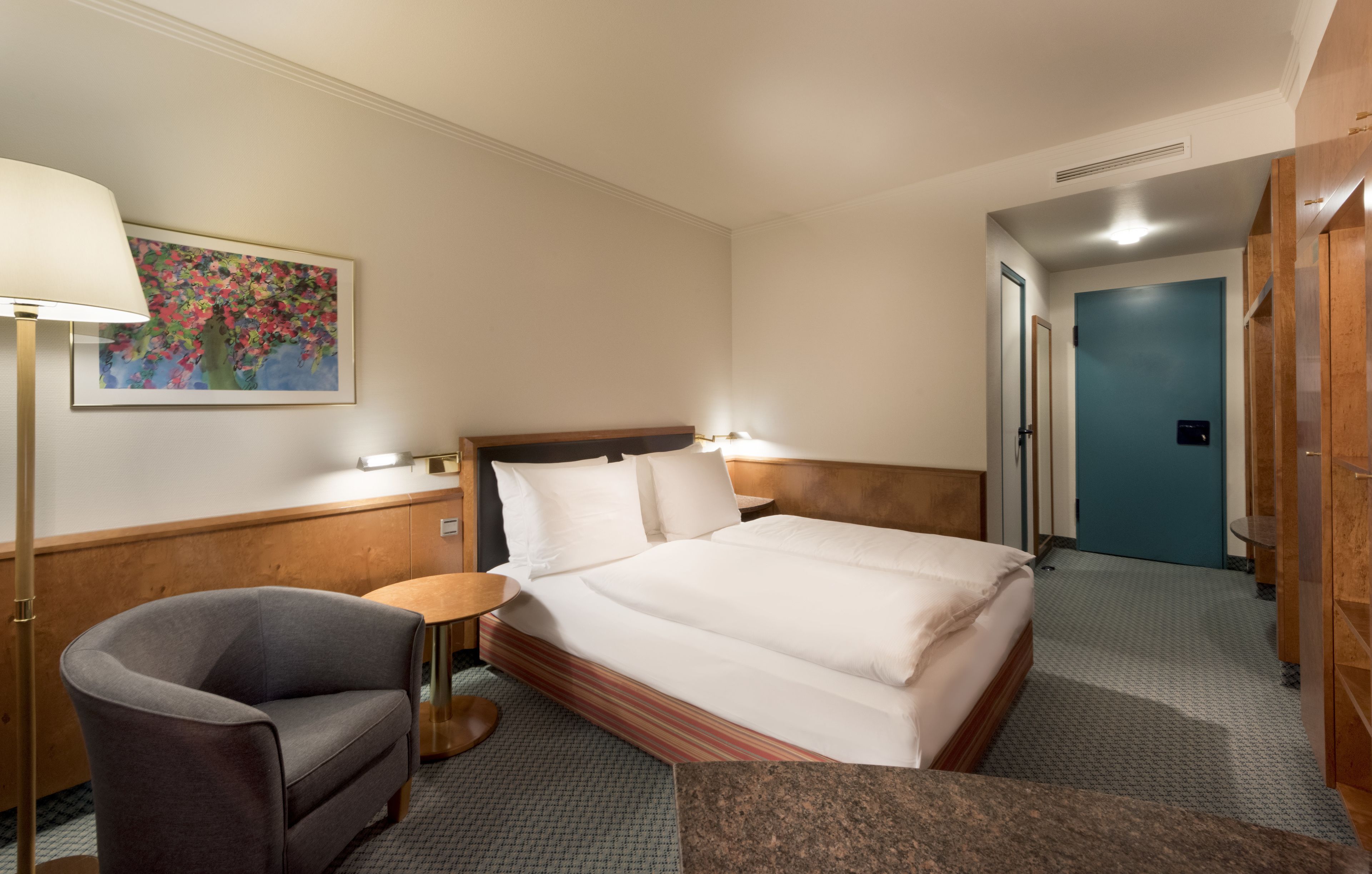 A hotel room with a bed, a chair, a small table, a painting on the wall, and a blue door in the background.