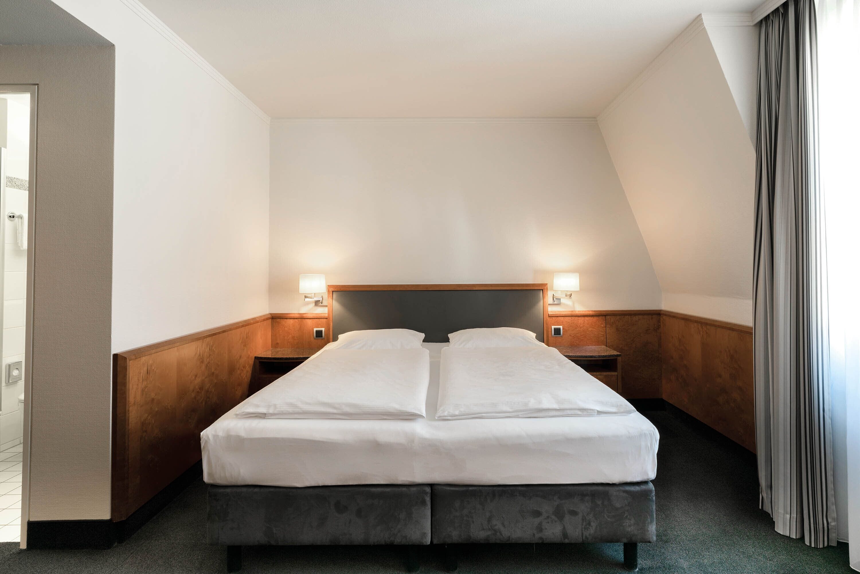 A hotel room with a double bed, two bedside tables with lamps, wooden paneling on the walls, and a view of the outside through a window with curtains.