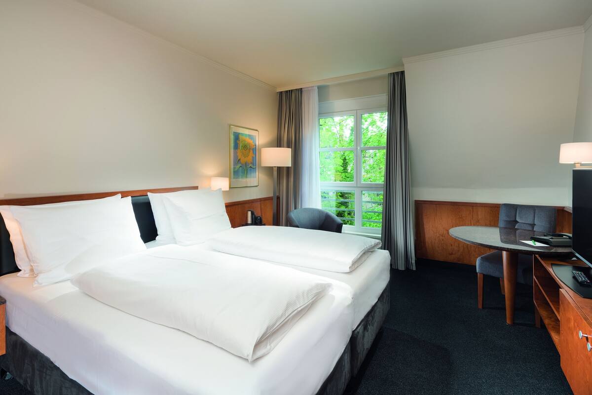 A hotel room with two beds, a desk, a chair, a TV, and a window with a view of greenery.