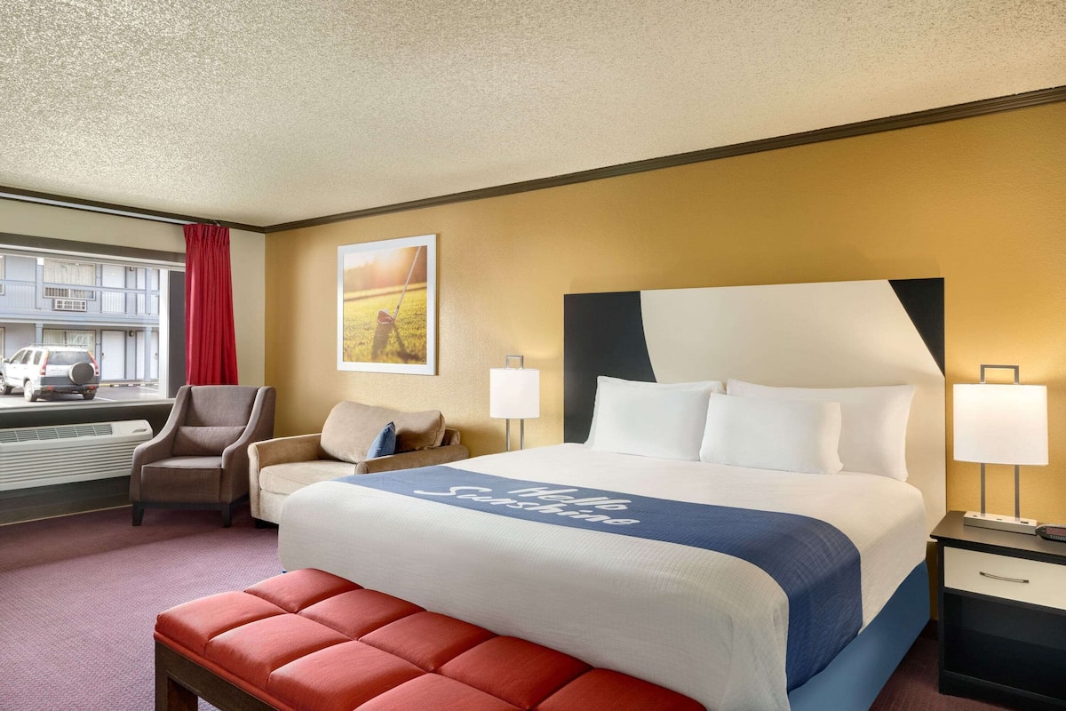A hotel room with a large bed, two bedside tables, a chair, a bench, a window with a view of a street, and a painting on the wall.