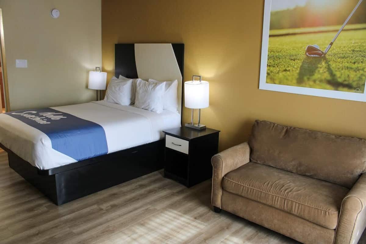 A hotel room with a bed, a nightstand, a lamp, a couch, and a wall-mounted picture.