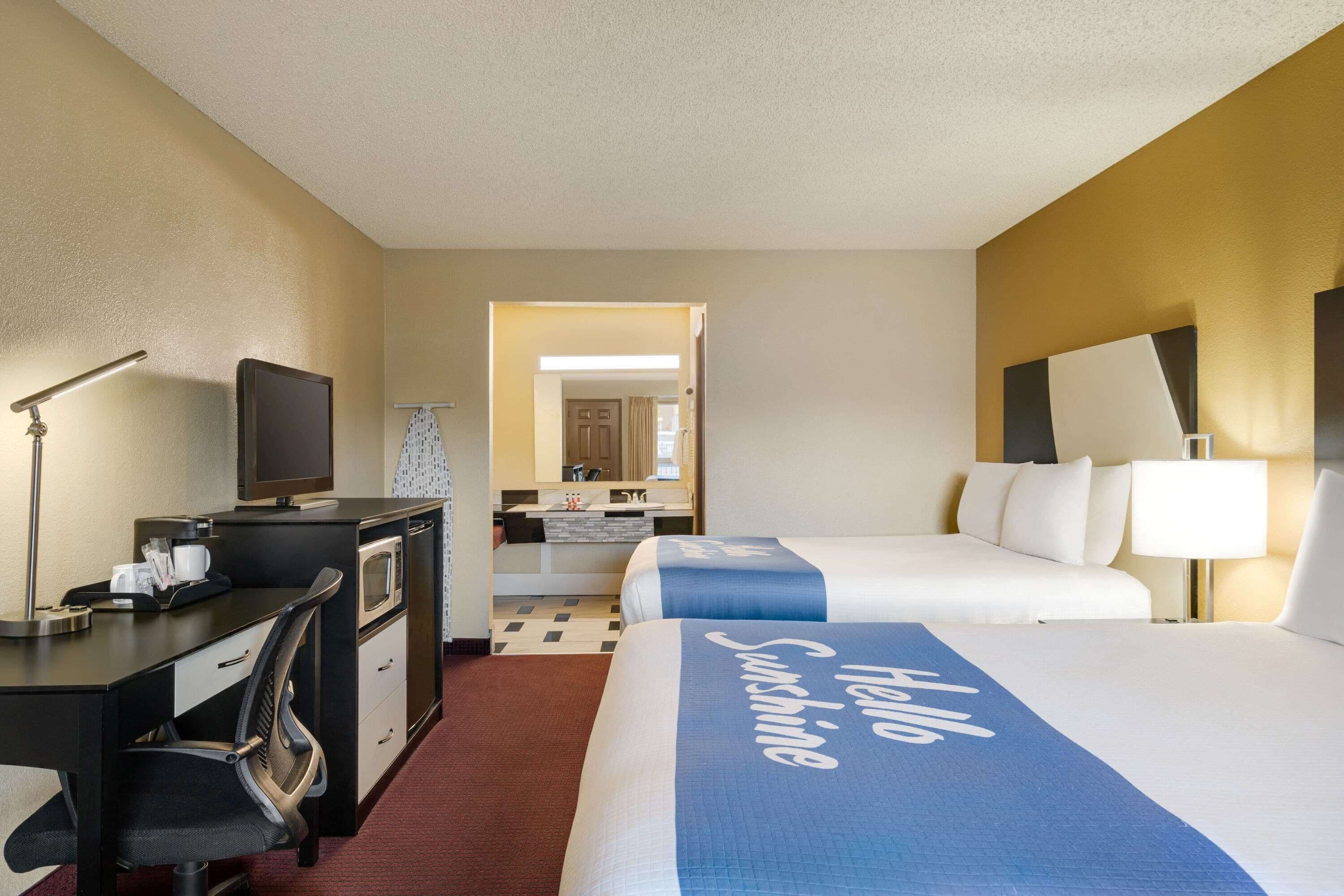 A hotel room with a desk, a television, a bed with a 'Hello Sunshine' bedspread, a chair, and a bathroom visible through an open door.