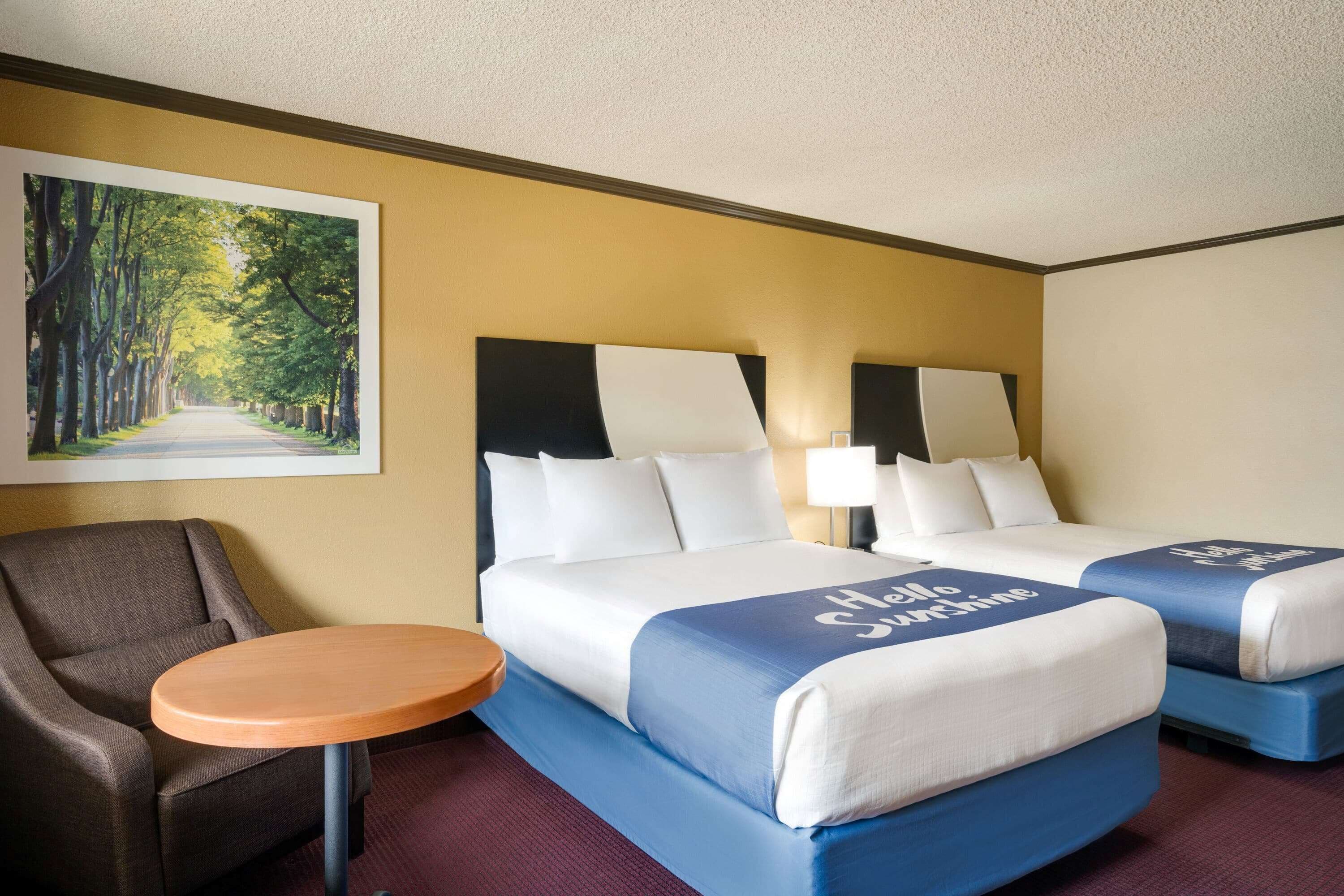 Ein Hotelzimmer mit zwei Betten, einem Sessel, einem kleinen Tisch und einem Bild einer von Bäumen gesäumten Straße an der Wand.