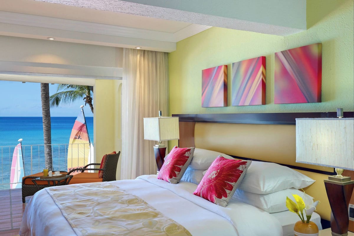 Une chambre d’hôtel avec un lit, des oreillers, une table de chevet, une lampe, un vase de fleurs et une vue sur l’océan.