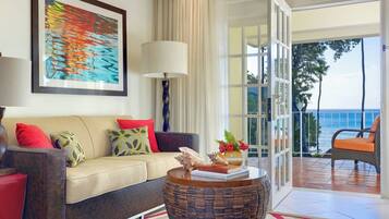 A living room with a beige sofa, red and yellow cushions, a wicker coffee table with books and shells, a large abstract painting, and a view of the ocean through a glass door.