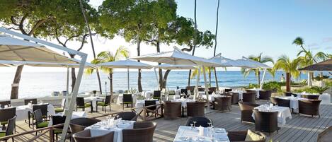 An outdoor dining area with tables and chairs, umbrellas, and a view of the ocean.