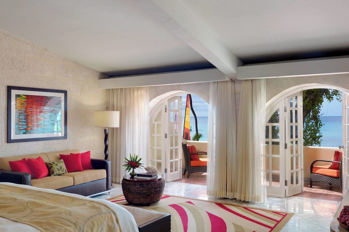 Une chambre d’hôtel avec un lit, un canapé, une table basse agrémentée d’une plante, et une vue sur la mer par une arche ouverte.