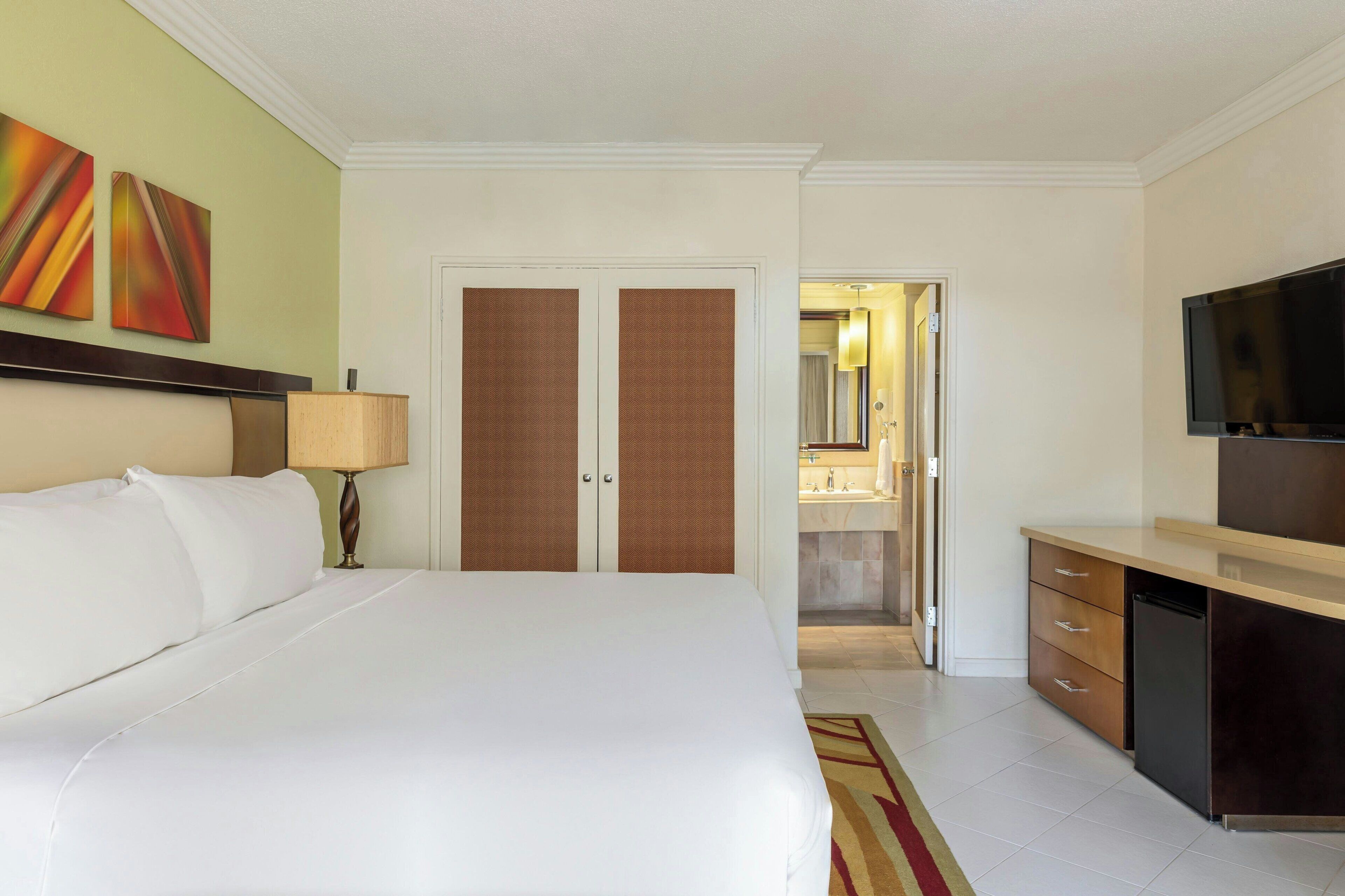A hotel room with a large bed, a desk with a television, a wardrobe, and a bathroom visible through an open door.