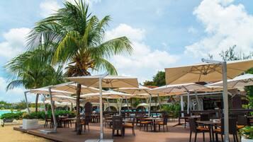 An outdoor dining area with umbrellas, tables, and chairs, overlooking a pool and beach.