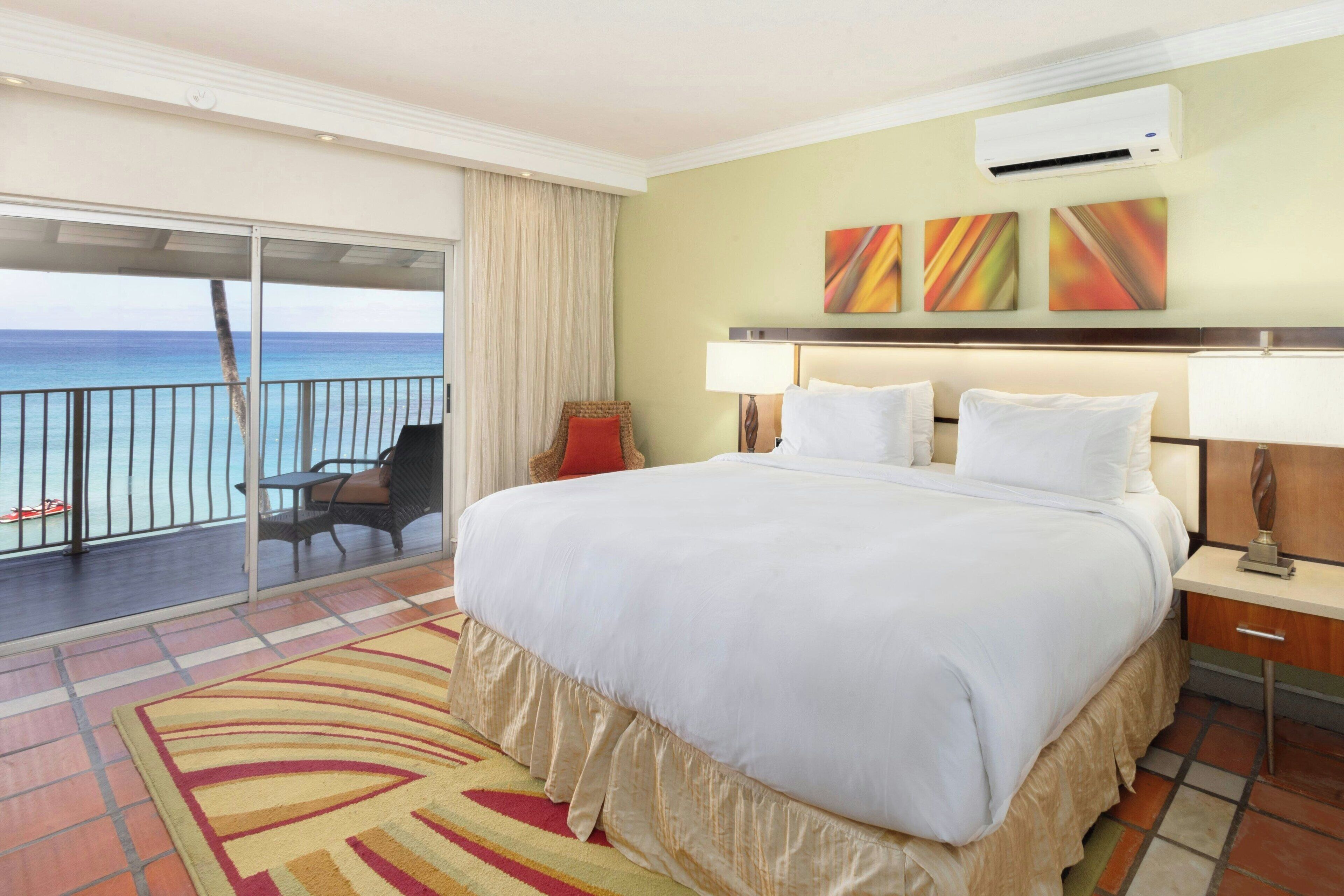 A hotel room with a large bed, a balcony with a view of the ocean, and a chair.