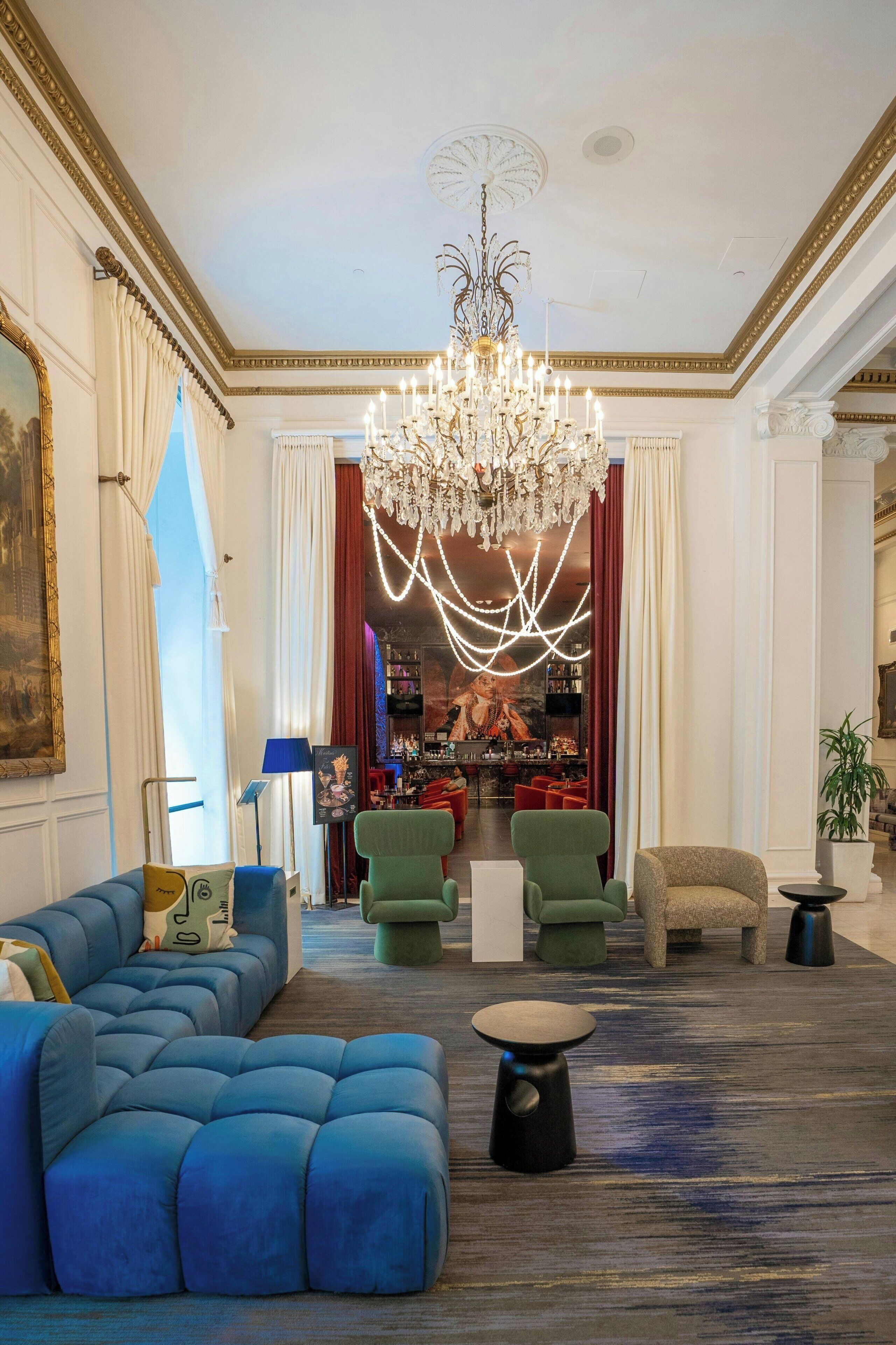 A spacious living room with a large blue sofa, green armchairs, a chandelier, and a bar area in the background.