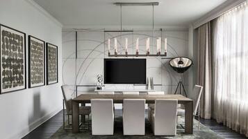 Une salle à manger moderne avec un grand téléviseur à écran plat, une table à manger en bois et quatre chaises. Les murs sont ornés d’œuvres d’art encadrées et une lampe sur pied se trouve dans la pièce.