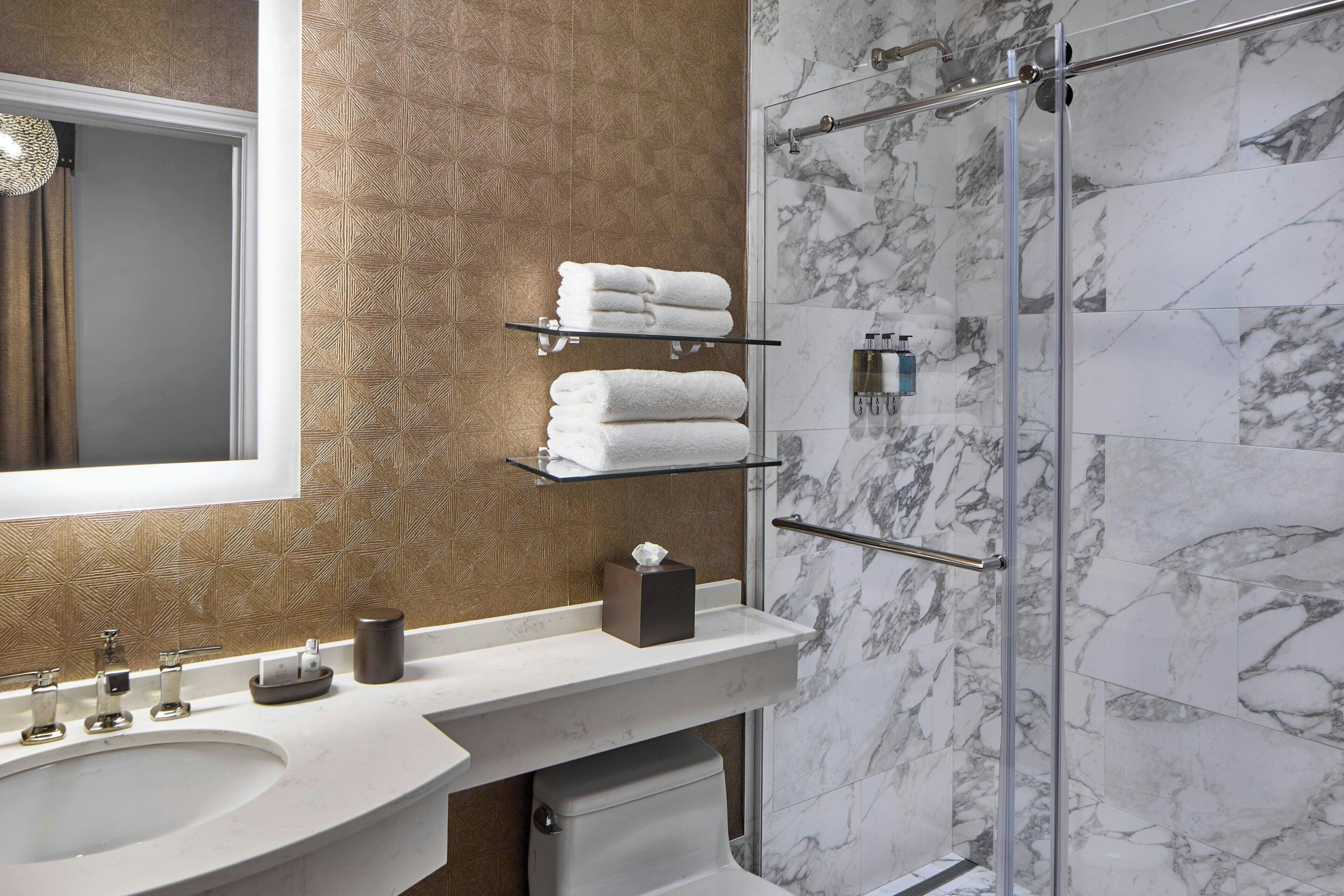 A modern bathroom with a marble shower, a glass shelf with towels, and a sink with a mirror above it.