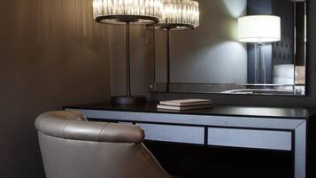 A modern hotel room with a dark-toned desk, a leather chair, a large mirror, and two chandeliers.