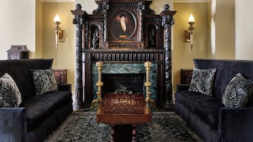 Une pièce avec deux canapés noirs, une table d’échecs en bois, une cheminée surmontée d’un portrait et des appliques murales.