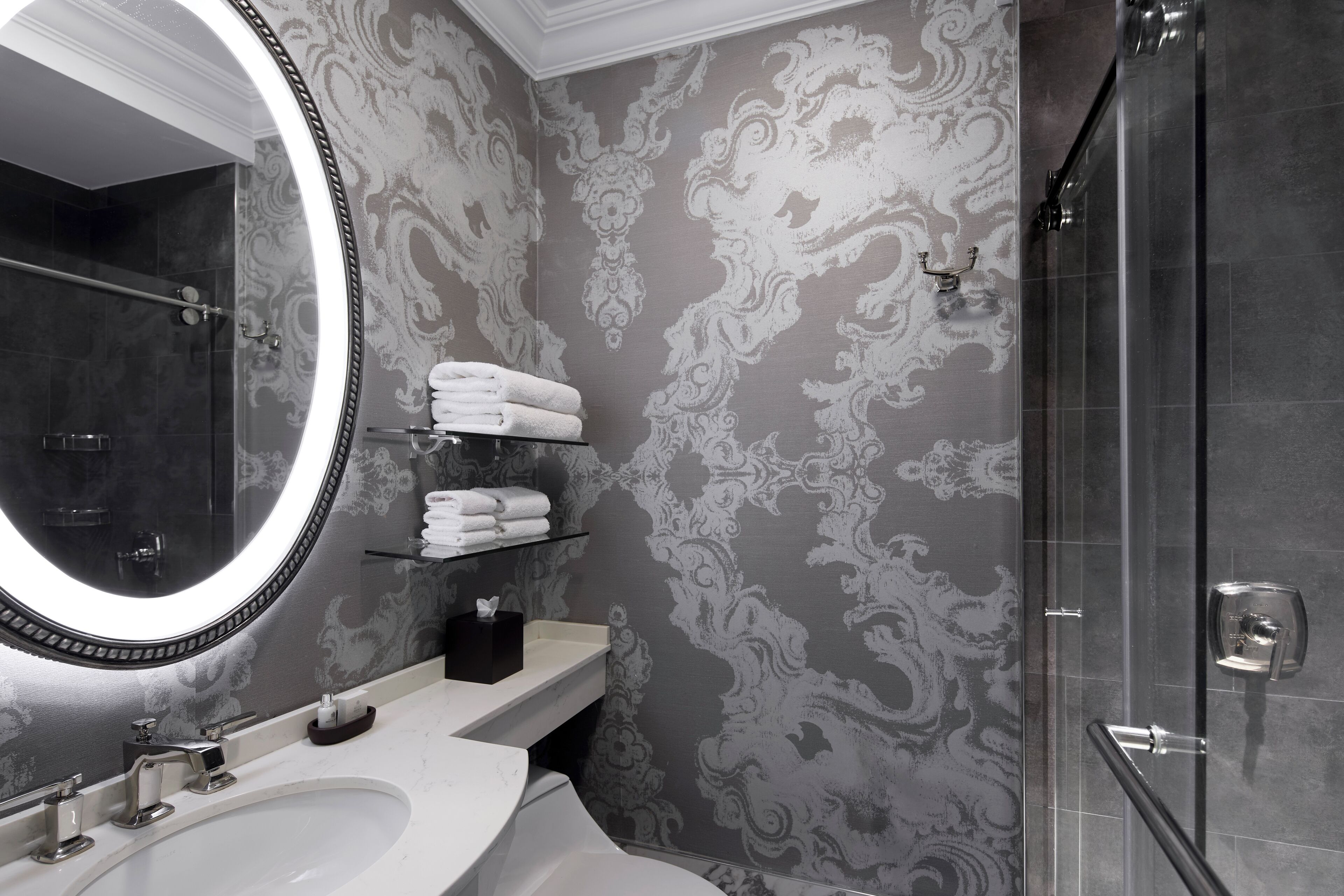 Une salle de bain dotée d’un grand miroir, d’un lavabo blanc, d’une cabine de douche en verre et d’un papier peint décoratif.