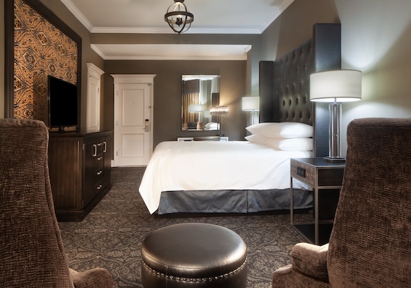 Quarto de hotel com cama, duas poltronas, escrivaninha com abajur, televisão e guarda-roupa.