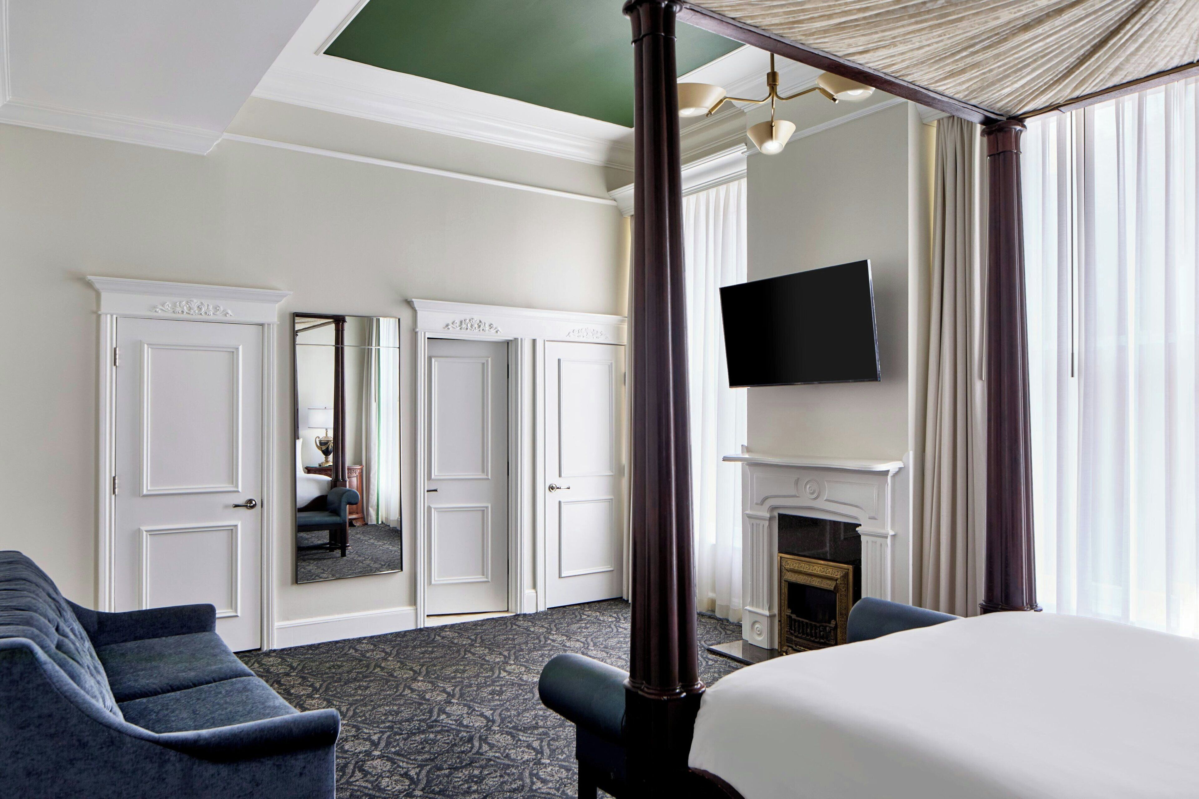 A hotel room with a four-poster bed, a flat-screen TV mounted above a fireplace, and a large mirror reflecting the room.