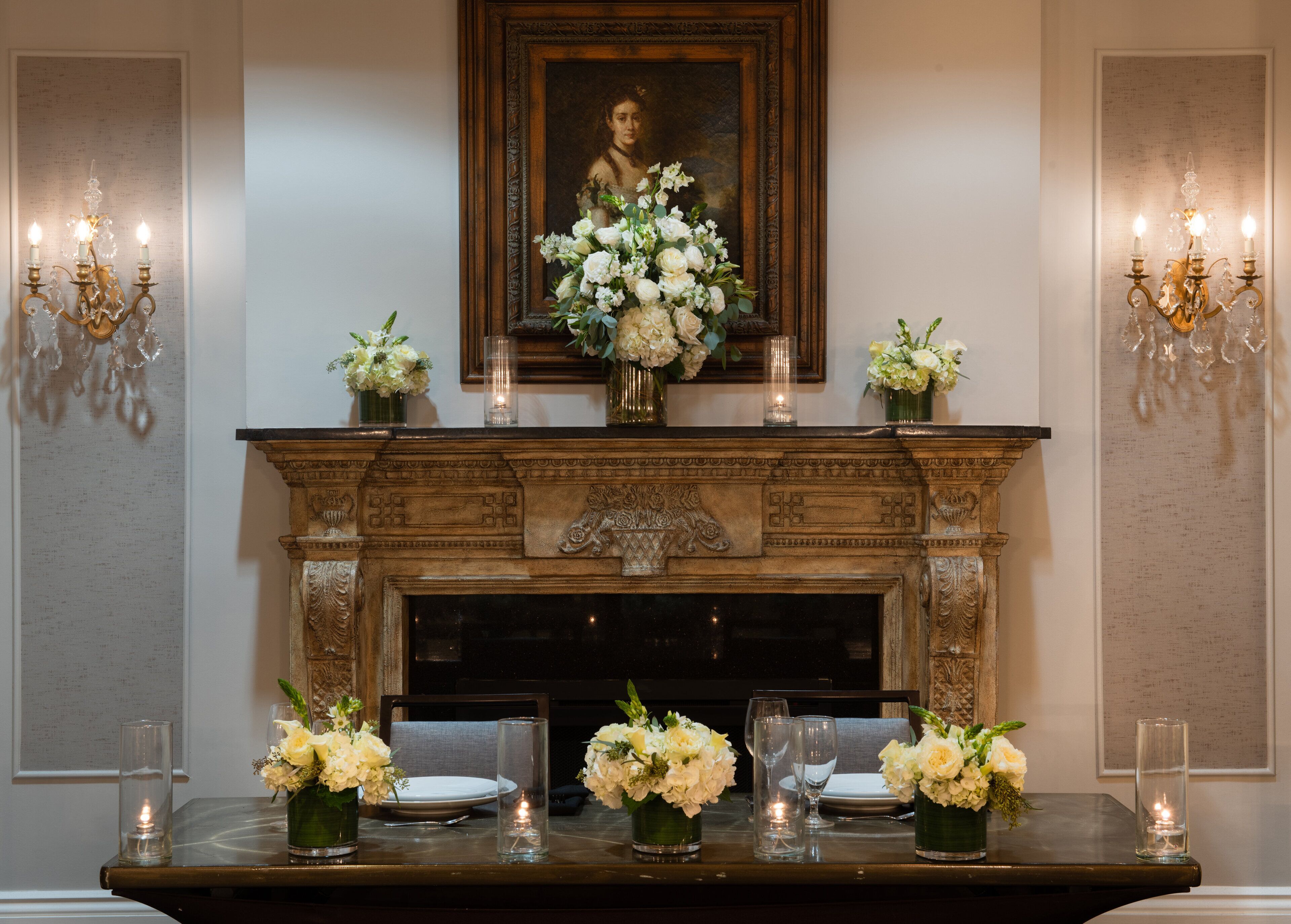 A formal dining room with a fireplace, a portrait above it, and candle-lit tables set for a meal.