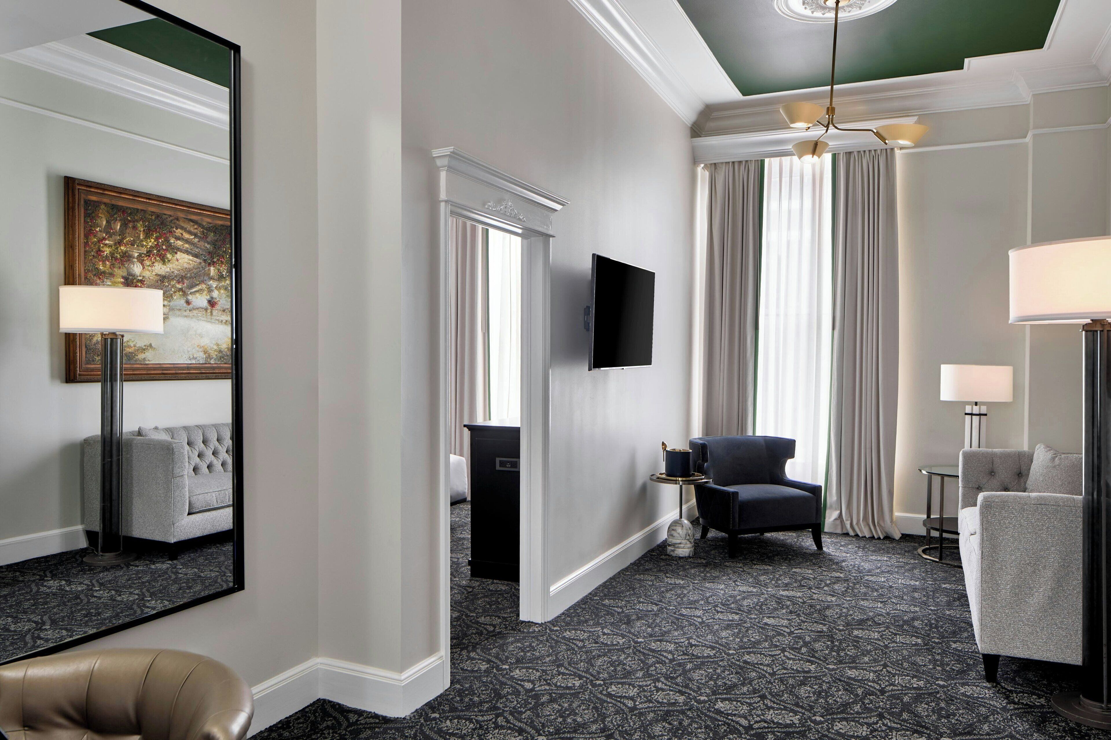 Une chambre d’hôtel dotée d’un grand miroir, d’un téléviseur à écran plat, d’un canapé, d’un fauteuil et d’une moquette à motifs.