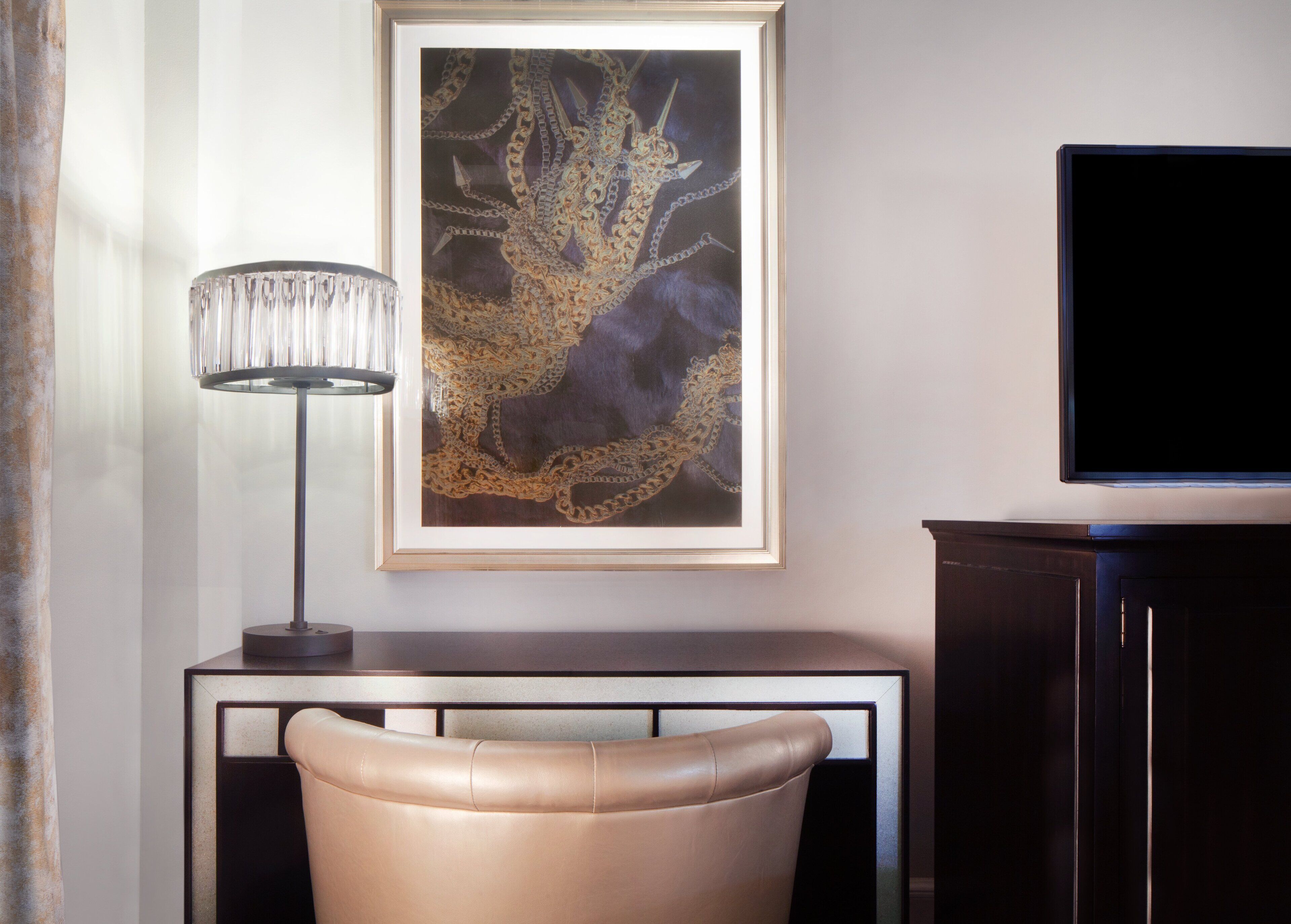 A framed artwork with a gold and black abstract design, a modern lamp, a leather chair, and a dark wooden cabinet with a flat-screen TV.