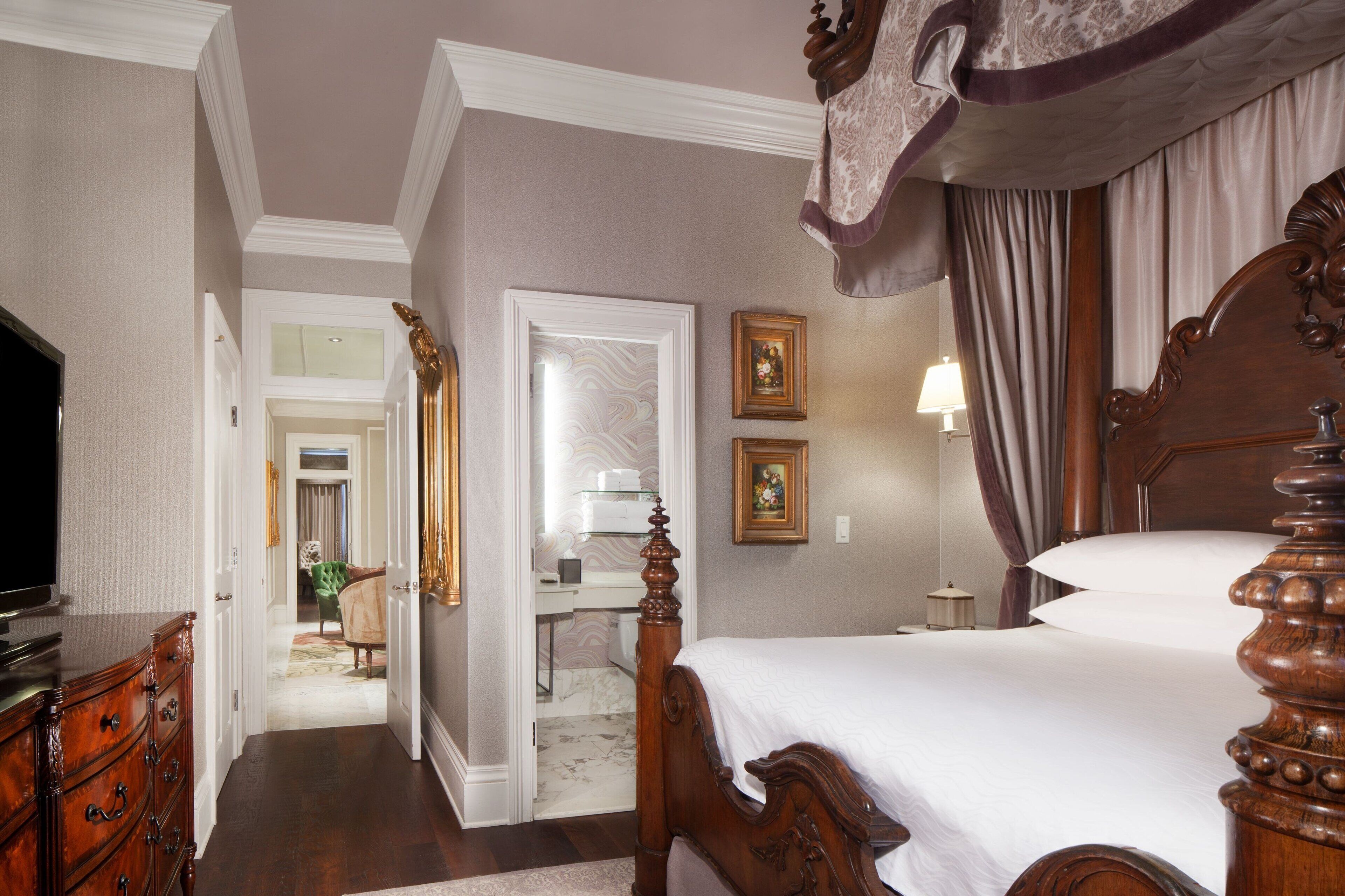 A bedroom with a large bed, wooden dresser, and a television. There is a bathroom visible in the background with a mirror and a sink.