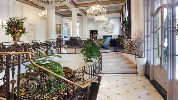 A grand lobby with ornate chandeliers, a marble staircase, and a seating area with a view of the city. - Le Pavillon, New Orleans, a Tribute Portfolio Hotel by Marriott (New Orleans)