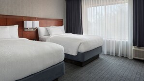 Une chambre d’hôtel avec deux lits, une tête de lit en bois, une table de chevet avec une lampe et une grande fenêtre avec des rideaux légers.