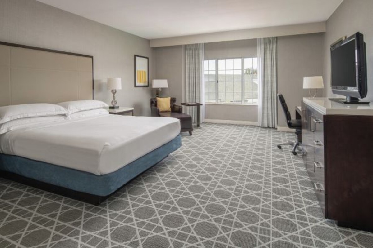 A hotel room with a large bed, a desk with a TV, a chair, a small table, and a window with curtains.