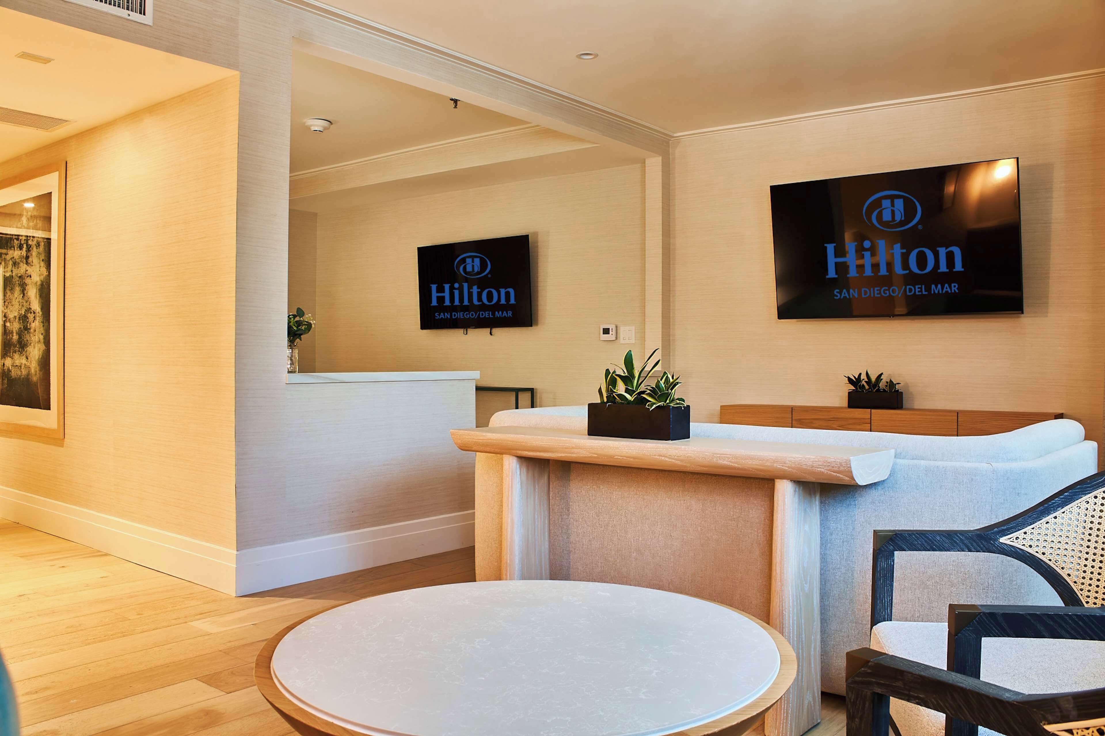 A modern hotel lobby with a reception desk, a flat-screen TV displaying the Hilton logo, and a seating area with a round table and chairs.