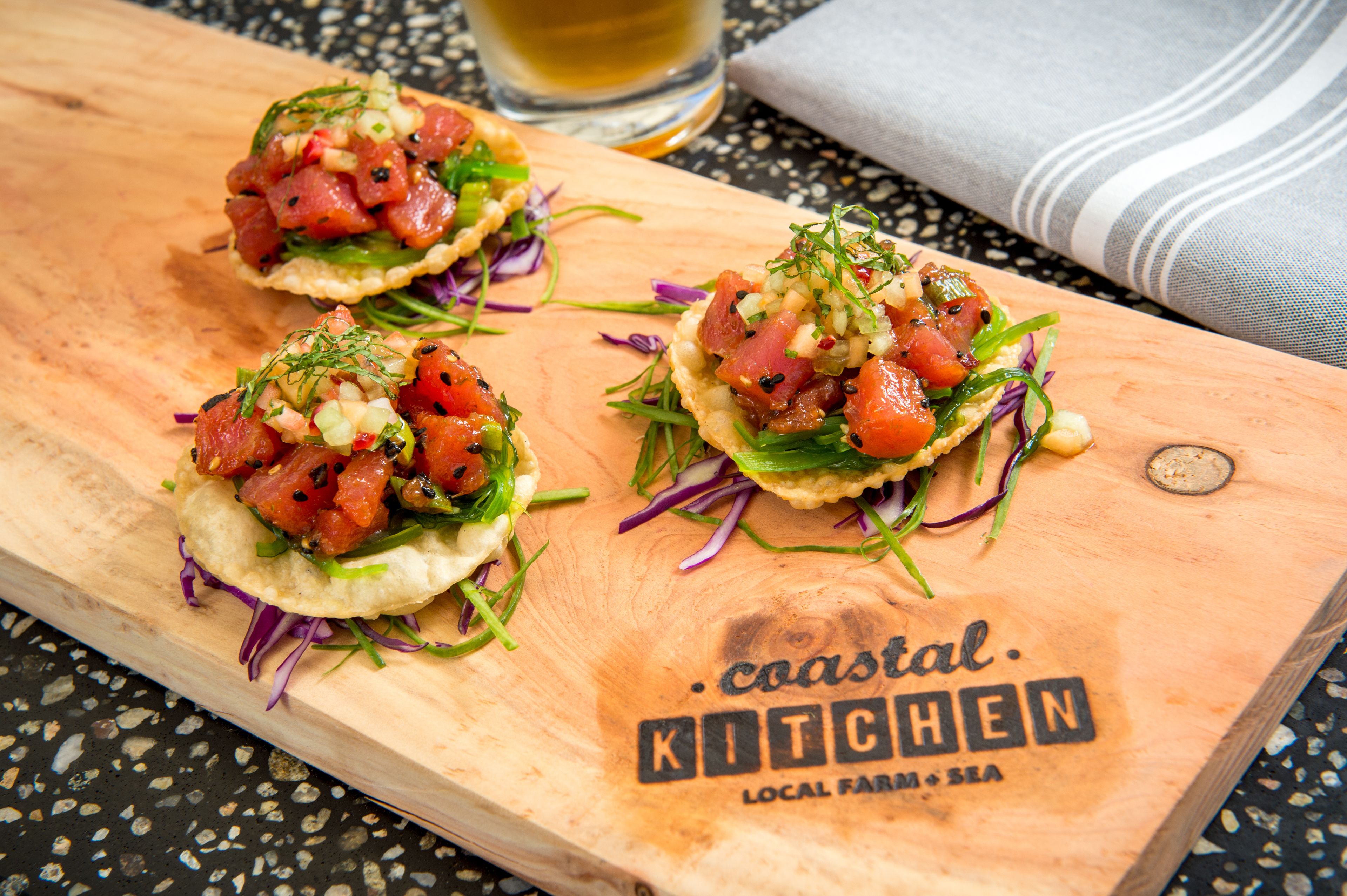 Three seafood appetizers on a wooden board with the text "coastal kitchen local farm & sea".