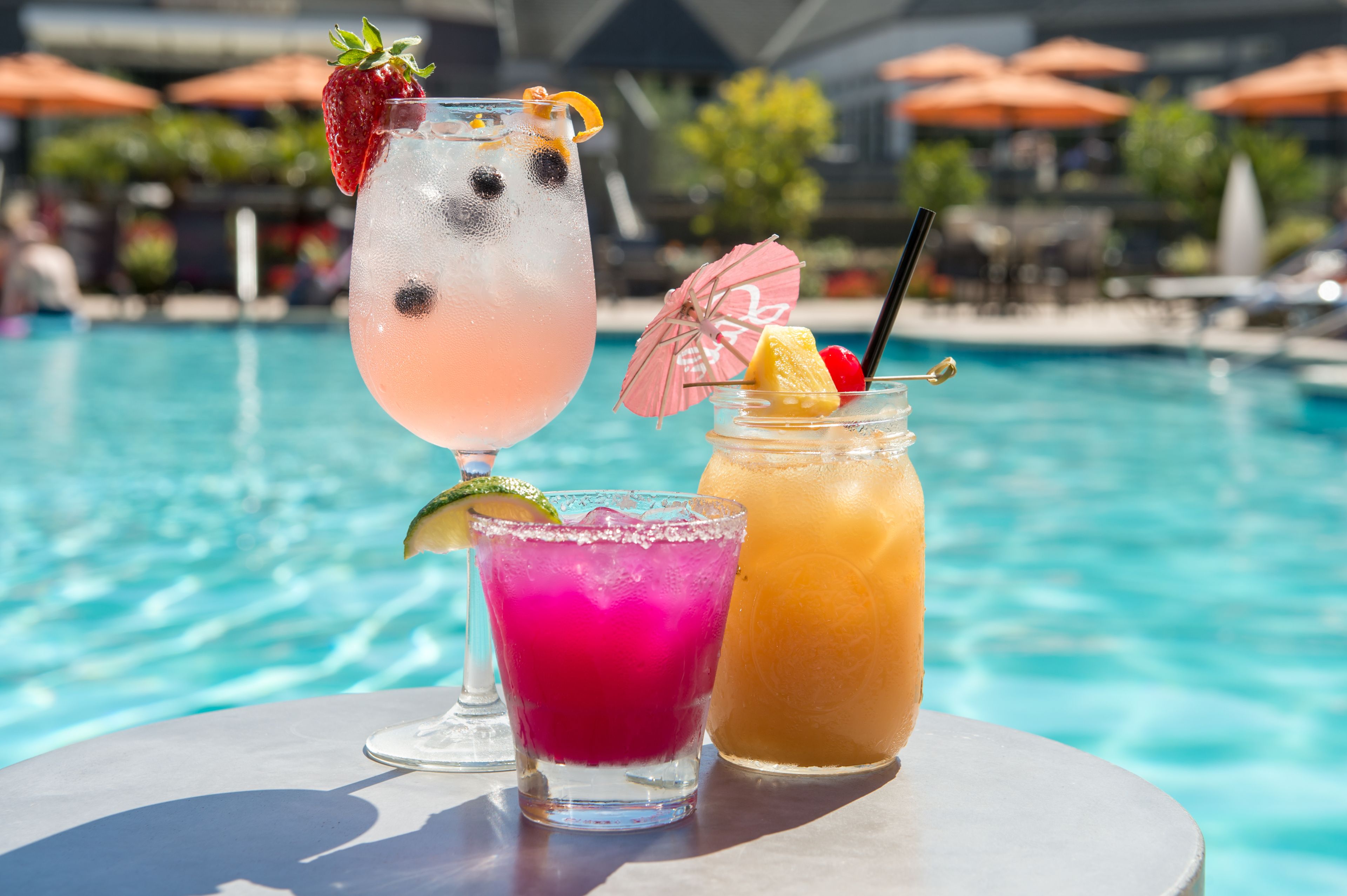 Tres bebidas sobre una mesa junto a una piscina, con sombrillas al fondo.