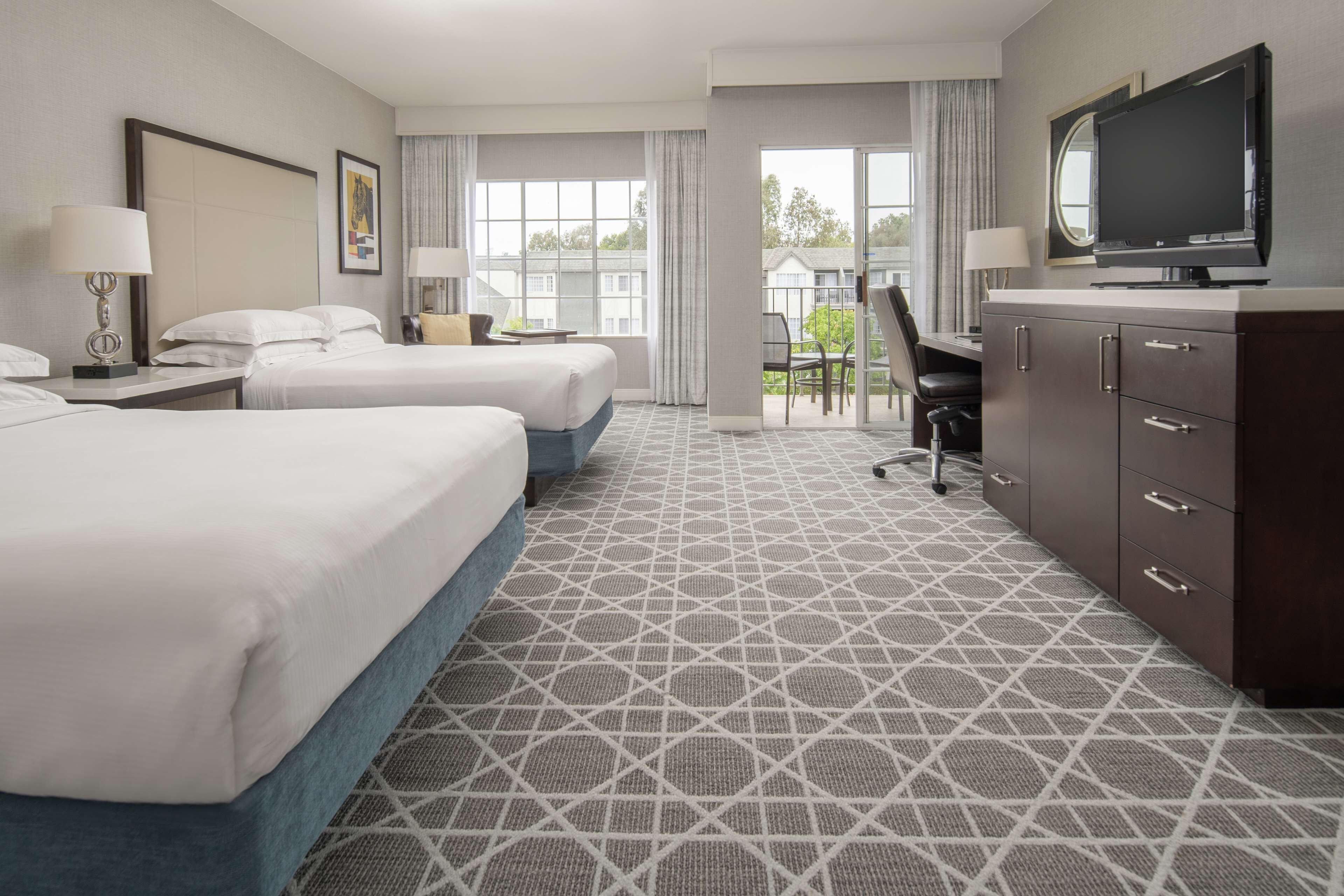 A hotel room with two beds, a desk, a television, and a view of the outside.