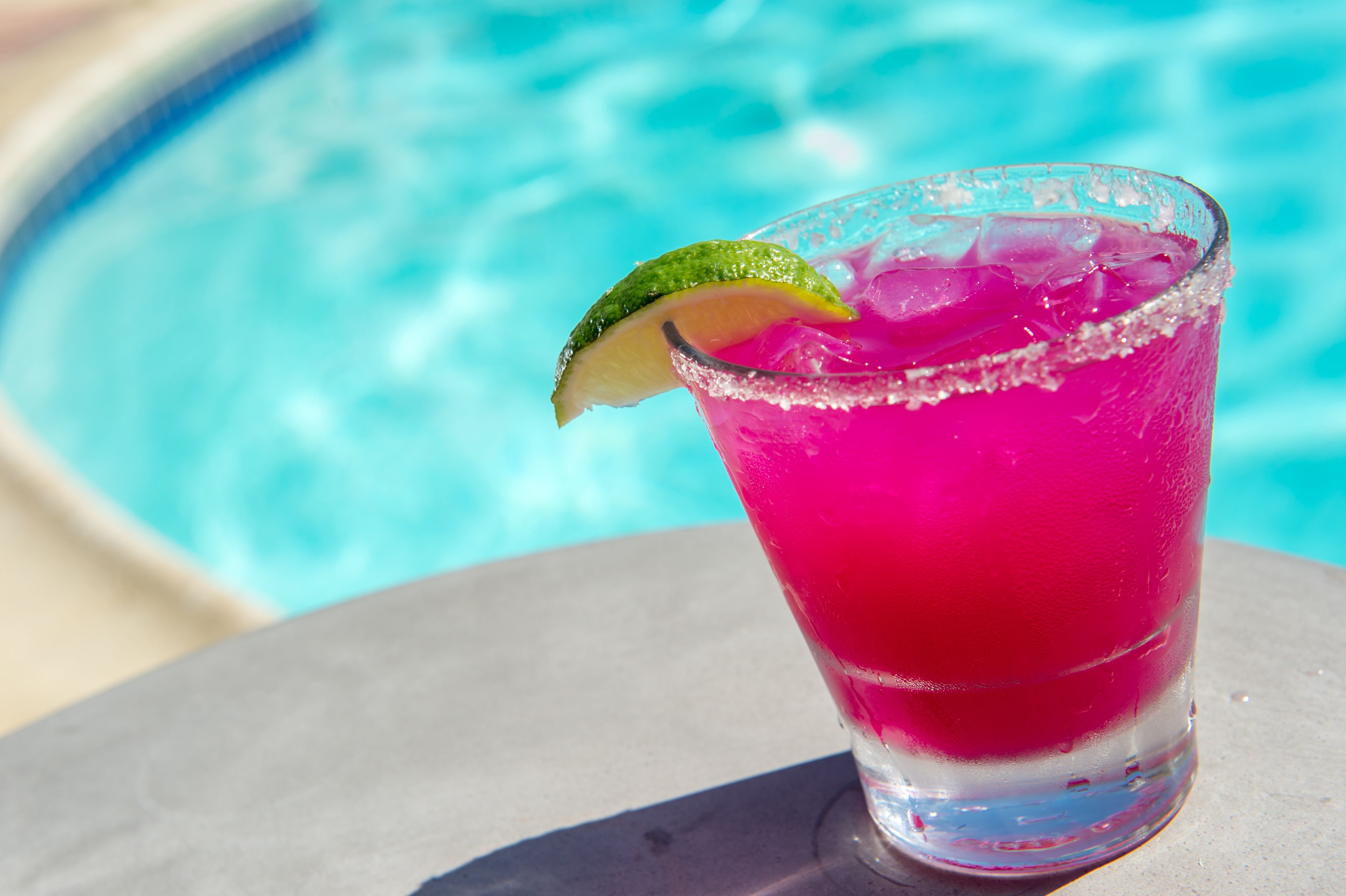 Un verre d’une boisson rose, garnie d’une tranche de citron vert, posé au bord d’une piscine.