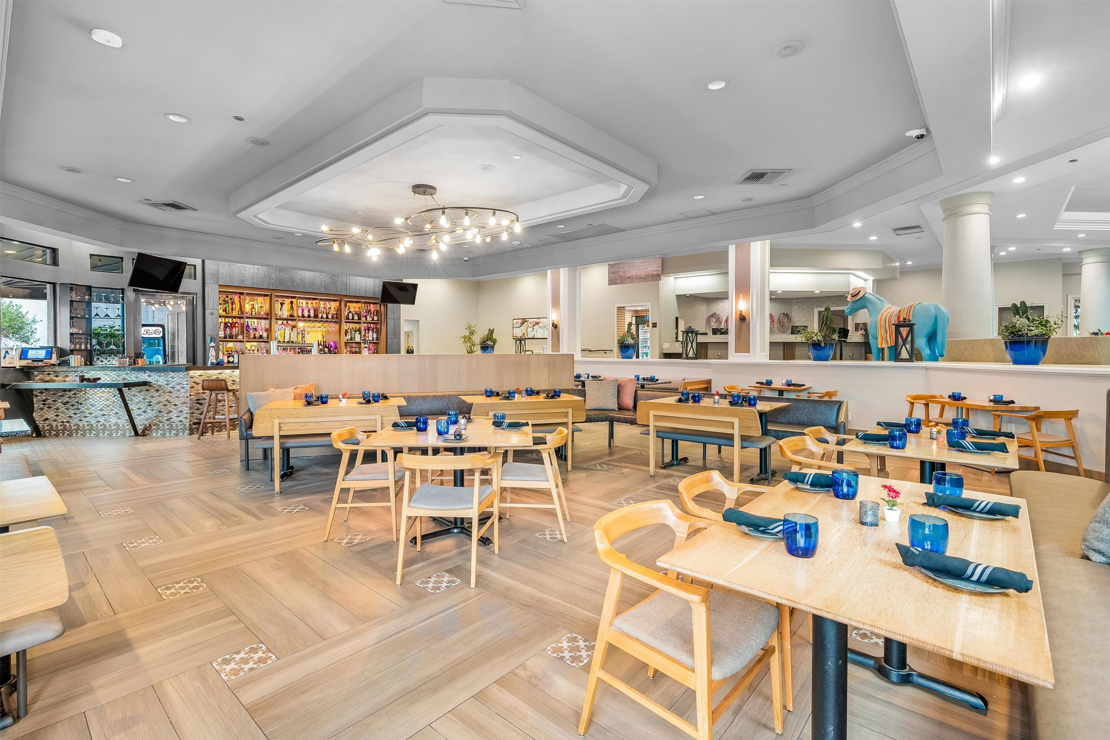 A modern restaurant with wooden tables and chairs, a bar area, and potted plants.