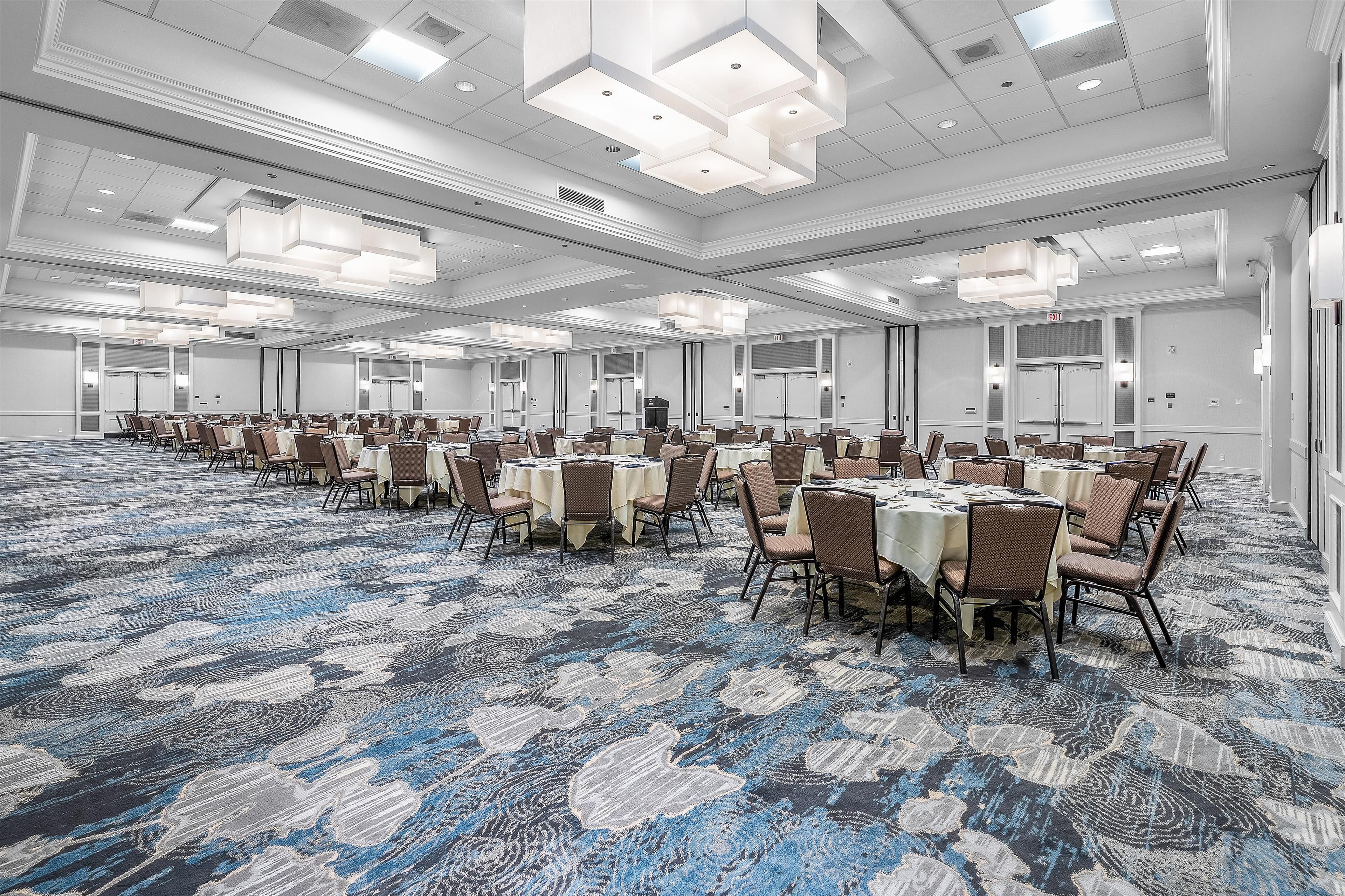 Une grande salle de conférence dotée de nombreuses tables et chaises, de hauts plafonds avec des luminaires encastrés et d’un tapis à motifs.
