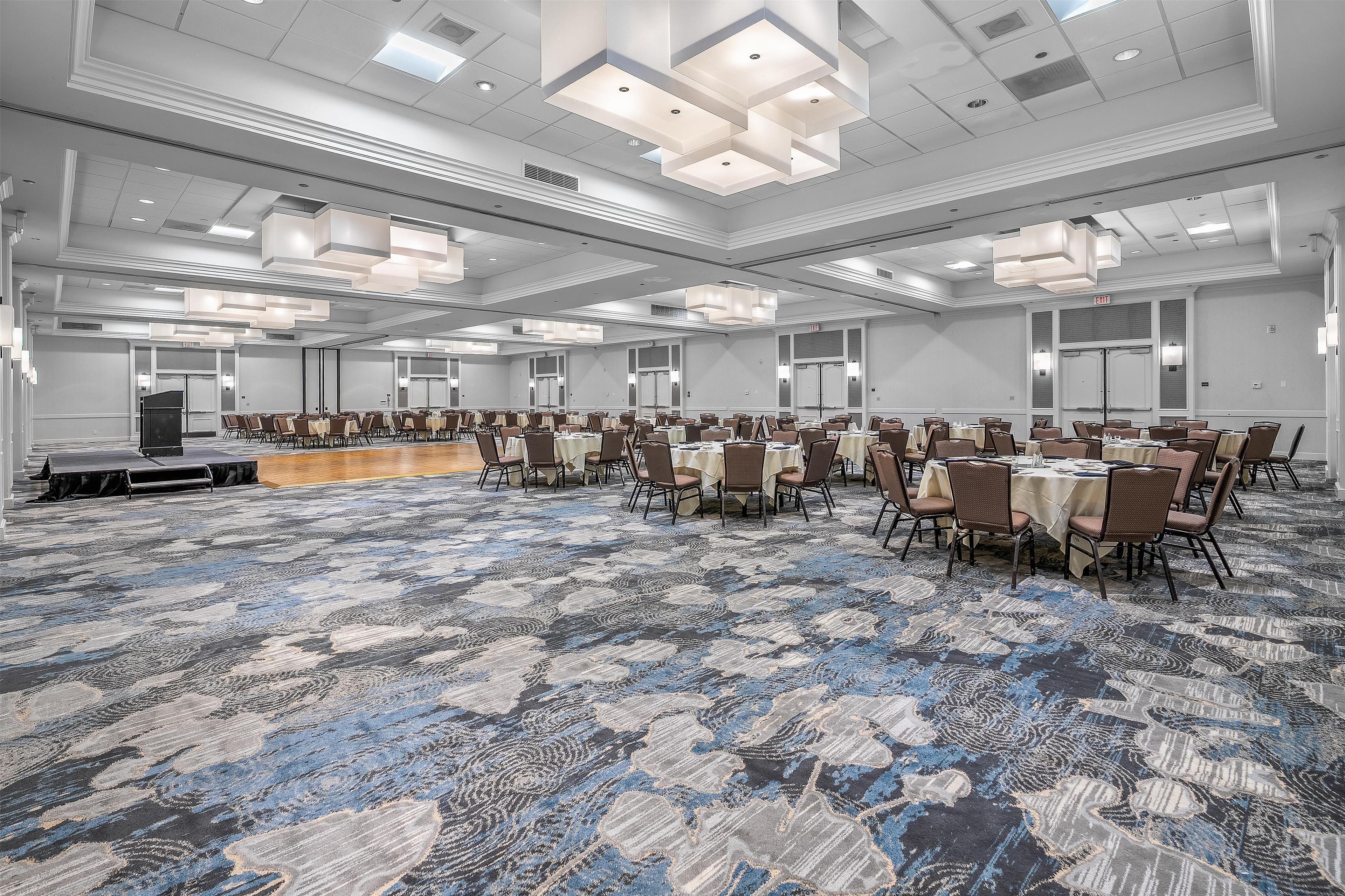 A large conference room with multiple tables and chairs, a stage, and a patterned carpet.