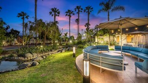 A well-lit outdoor patio area with a curved seating arrangement, surrounded by palm trees and a small water feature.