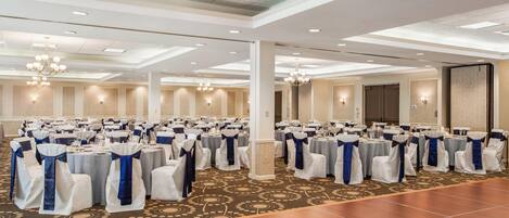 A large banquet hall with rows of round tables, white chairs with blue sashes, and a patterned carpet.