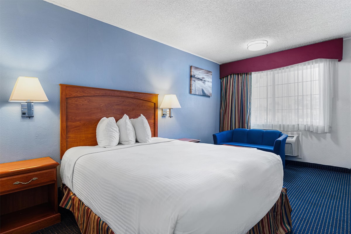 A hotel room with a large bed, two bedside lamps, a blue armchair, a window with curtains, and a painting on the wall.