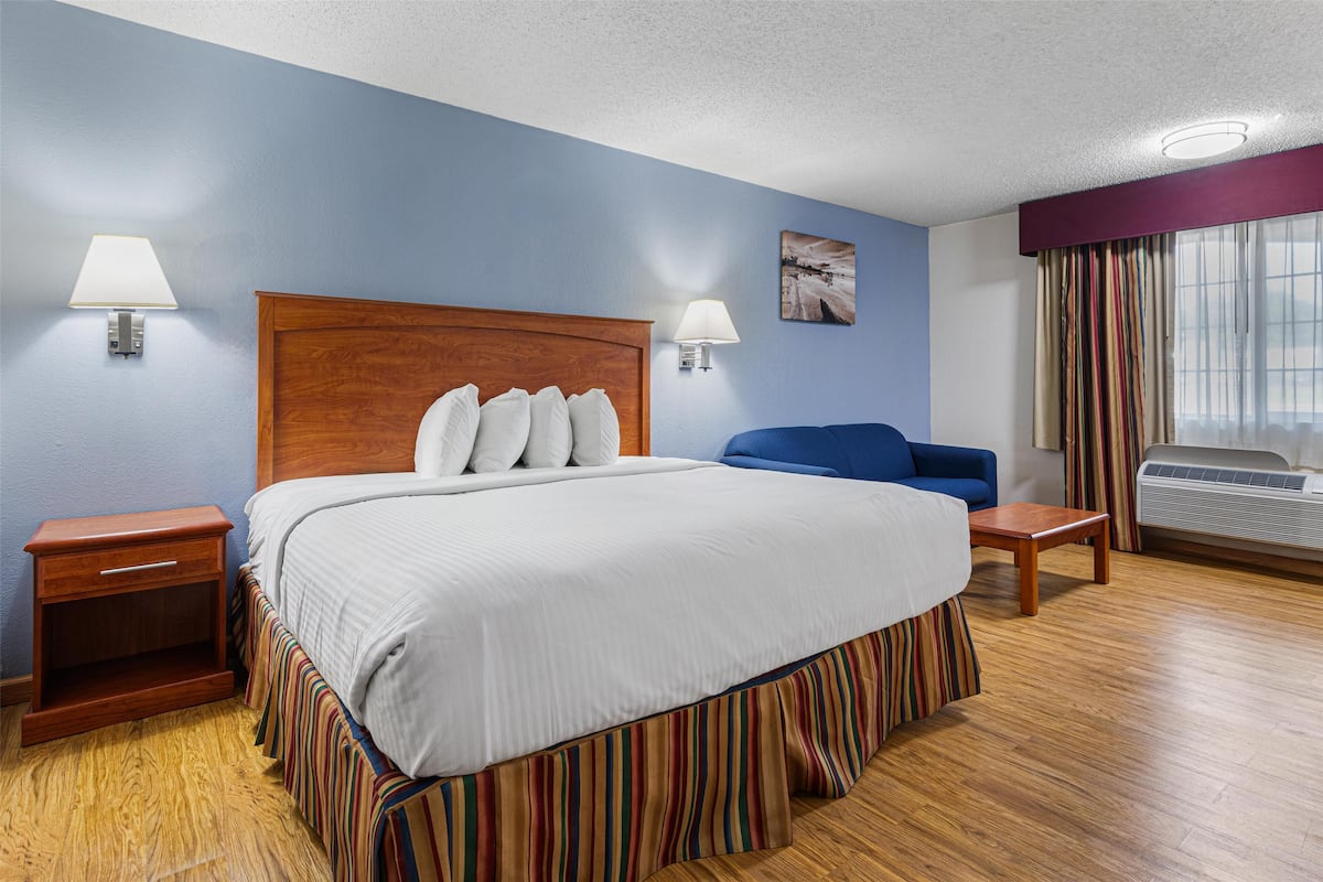 A hotel room with a large bed, two bedside tables, a blue sofa, a window with curtains, and an air conditioning unit.