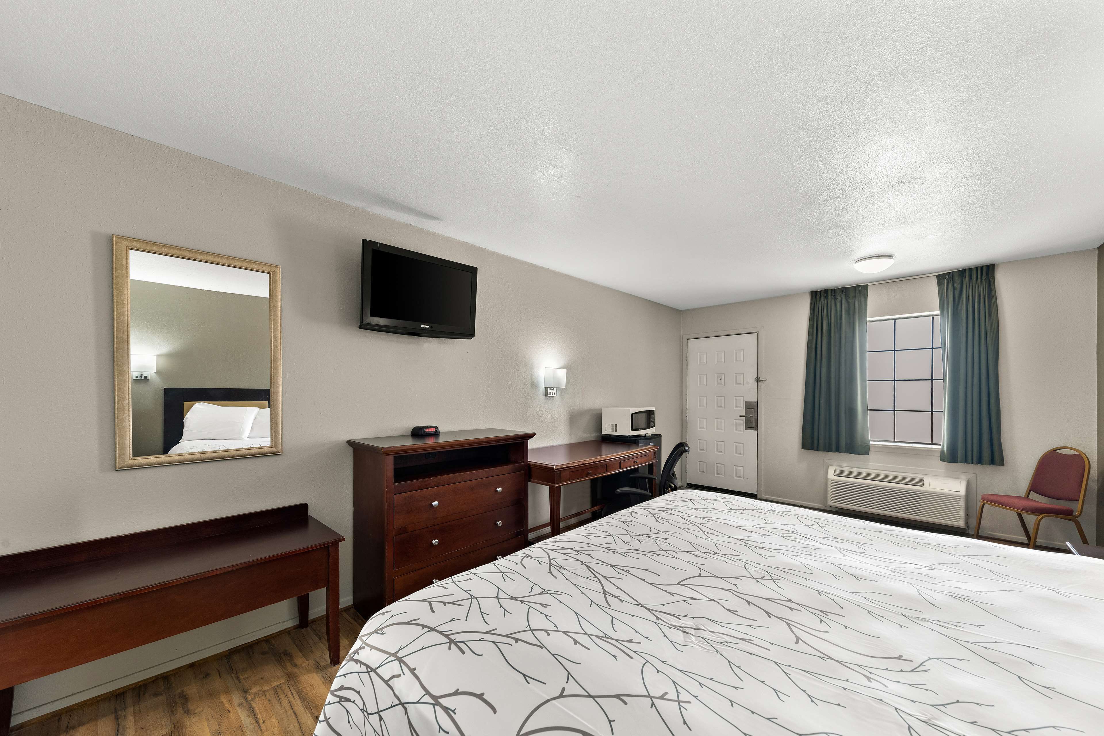 A hotel room with a large bed, a wooden dresser, a television mounted on the wall, a desk with a chair, and a window with curtains.