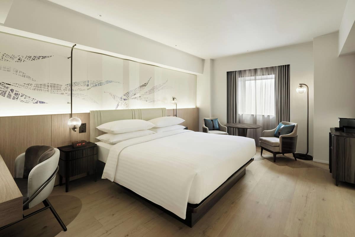 A modern hotel room with a large bed, a desk, a chair, a small table, a lamp, and a large window with curtains.