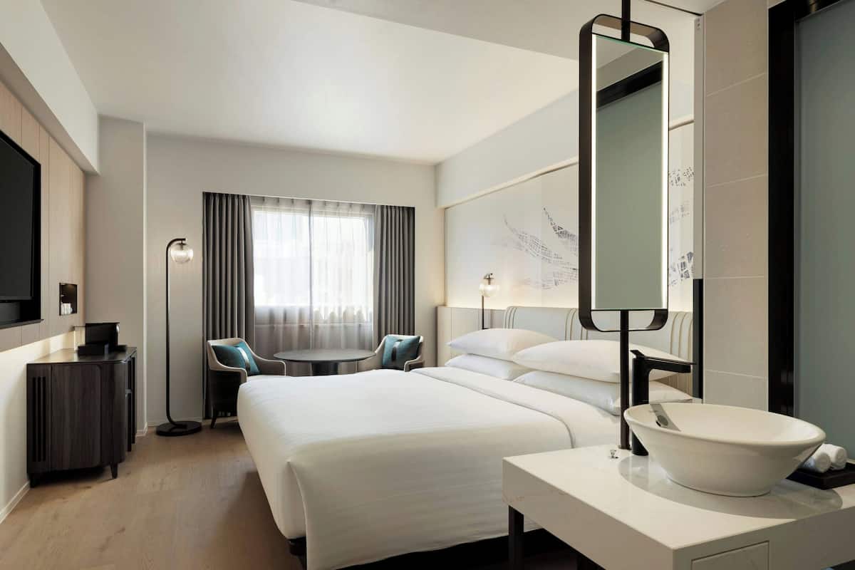 A hotel room with a large bed, a round table, a desk, and a modern bathroom with a large mirror and sink.