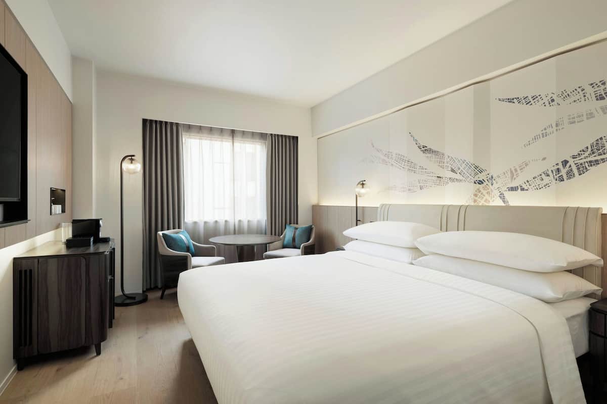 A hotel room with a large bed, a desk, a chair, a small table, and a large window with curtains.