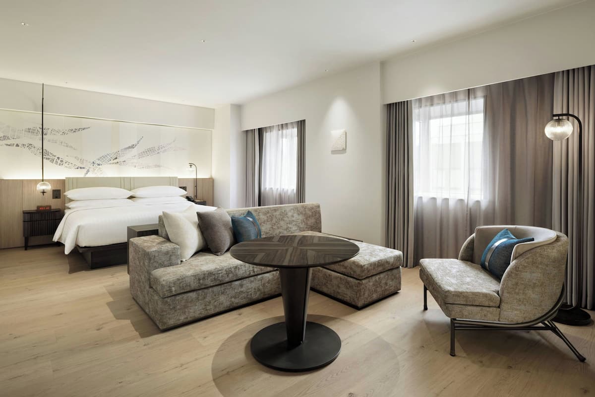 A modern hotel room with a bed, a sofa, a chair, a round table, and a large window with curtains.