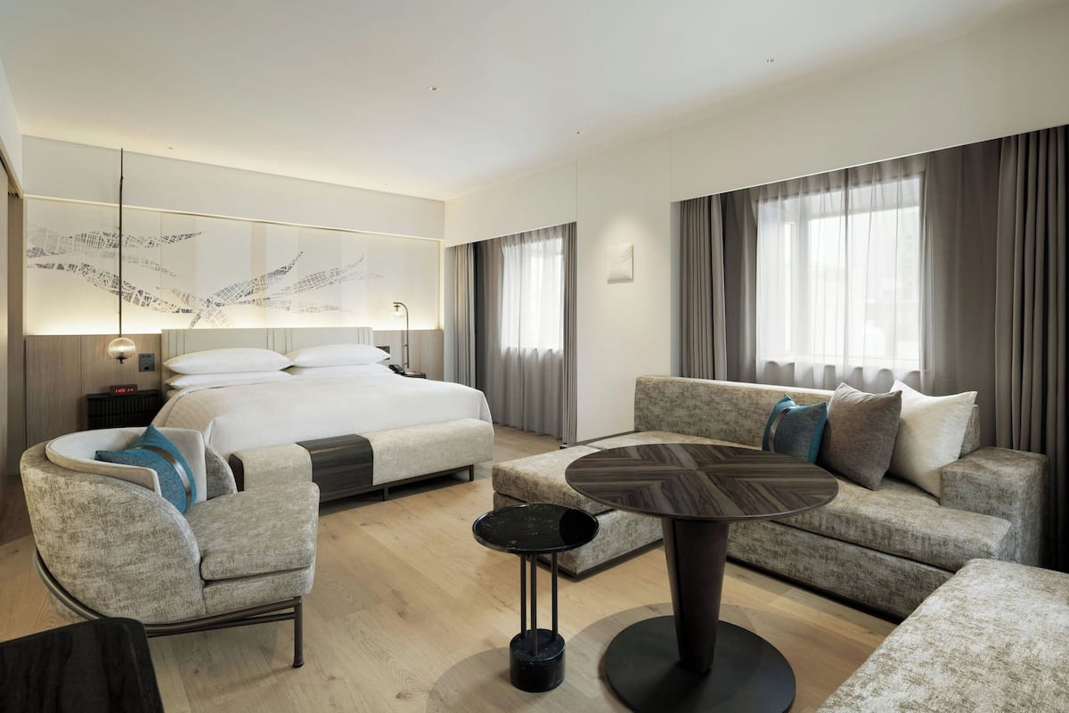 A modern hotel room with a large bed, a sofa, a round coffee table, and a wall-mounted artwork.