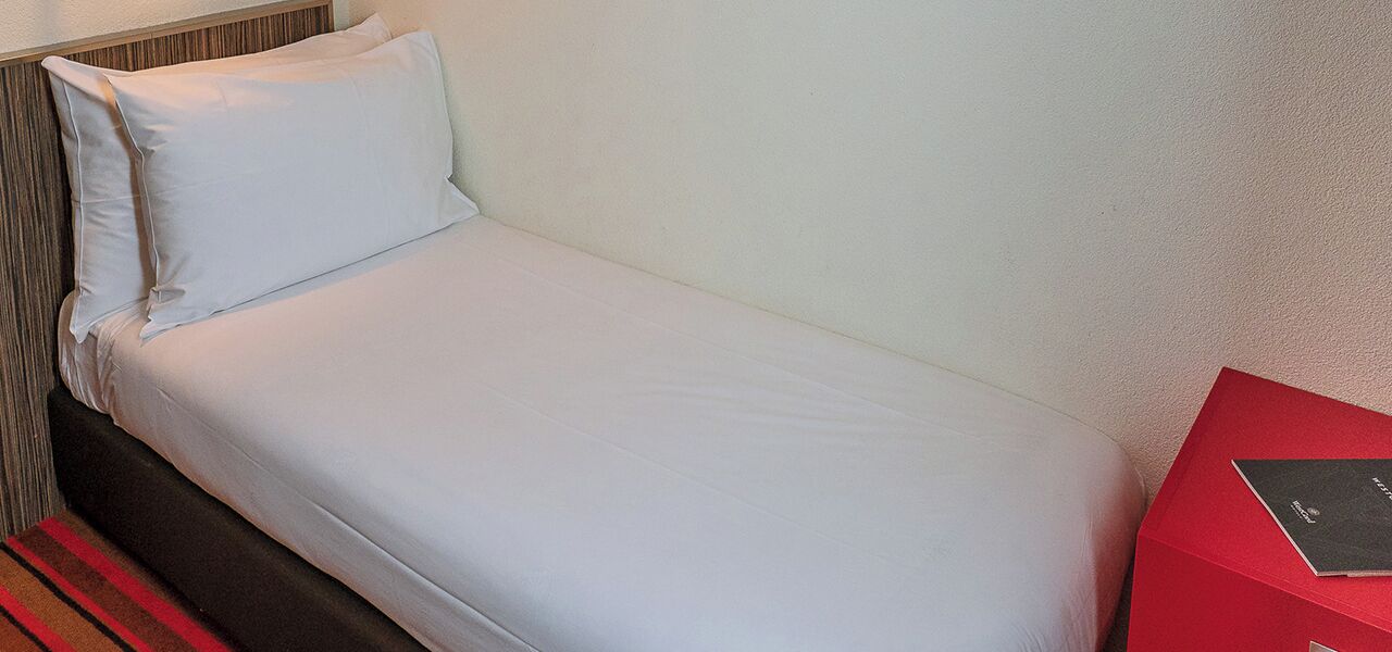 Un lit simple avec un matelas et un oreiller blancs, une tête de lit en bois et un objet rouge sur le côté droit.