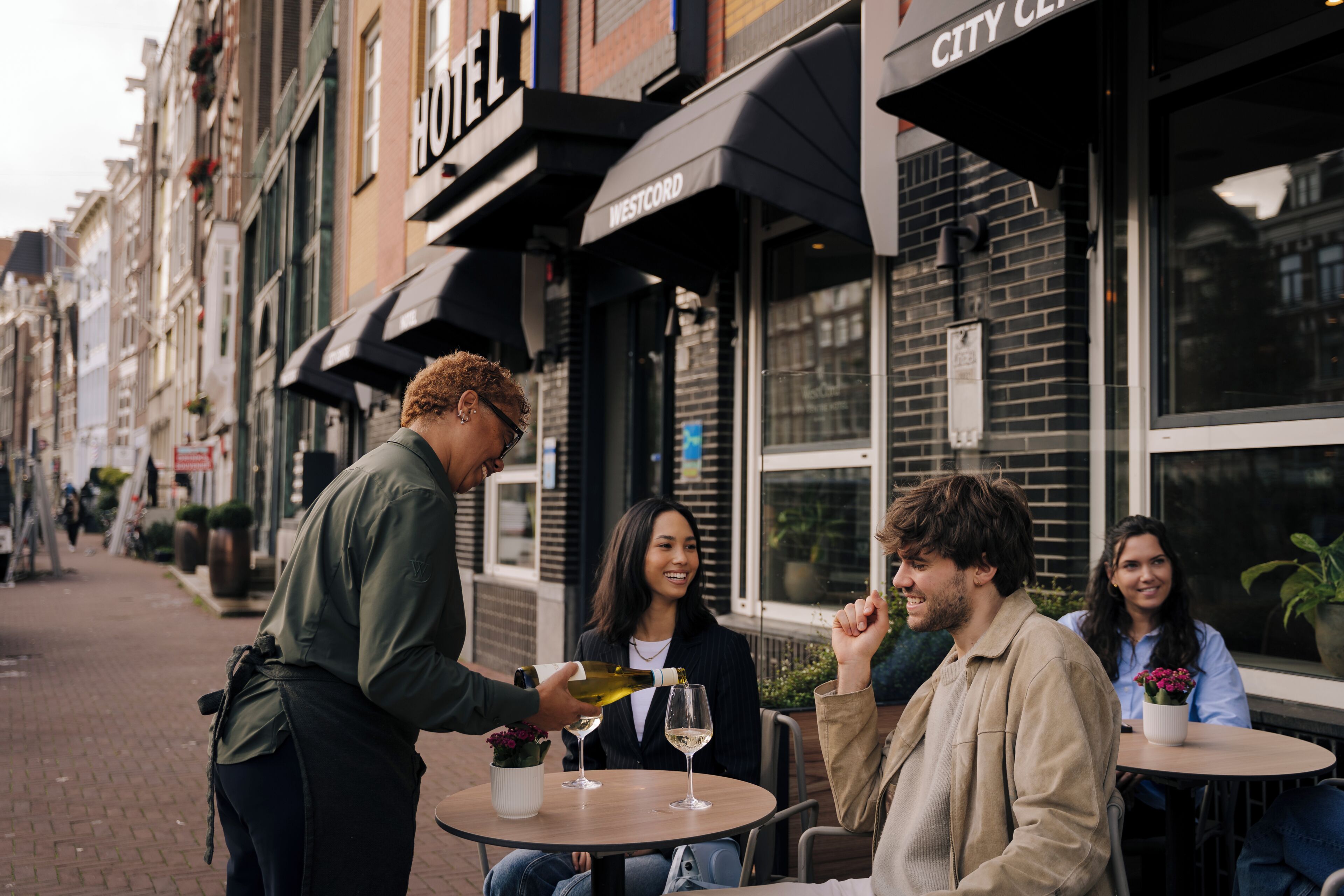 Escena callejera con un hotel llamado 'City Center' y una persona sirviendo vino a clientes en una mesa al aire libre.