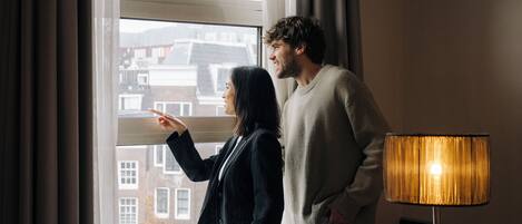 A room with a large window, a standing lamp, and a view of buildings outside.